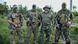 A group of four men wearing military fatigues, standing with weapons in a grassy field, looking at the camera