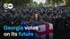 The photo shows a woman who has tied the flags of Georgia and the EU together and is holding them up. People dressed in black who are wearing masks are standing behind her. The photo is captioned "Georgia votes on its future".